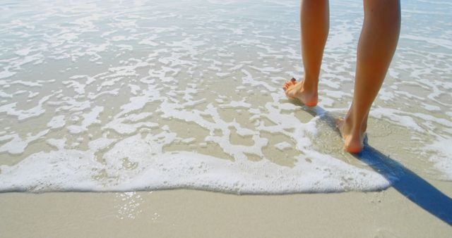 Bare feet of a person walking on a sandy beach while gentle ocean waves lap against the shore. Perfect for advertisements about travel destinations, oceanfront resorts, summer vacations, and nature retreats.
