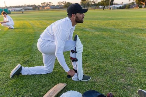 Kneeling cricket player fastening leg pad wearing white kit and black cap at golden hour