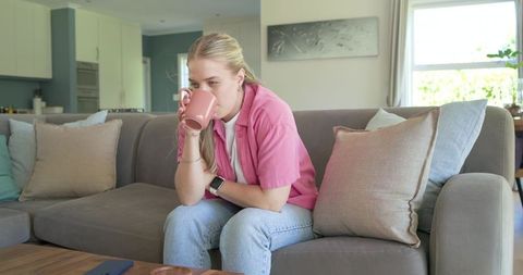 Pensive young woman sipping coffee on modern gray sofa with smartwatch on wrist, casual lifestyle
