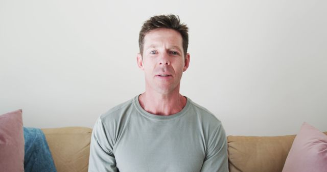 Mid-adult man sitting on a couch at home, looking directly at the camera. He appears calm and is wearing casual clothing, perfect for lifestyle, home life, and relaxation scenes. Ideal for use in articles or advertisements related to home living, daily routines, and men's fashion.