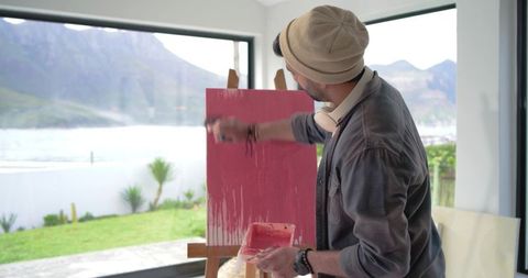 Indian artist painting red abstract canvas by waterfront window with mountain view