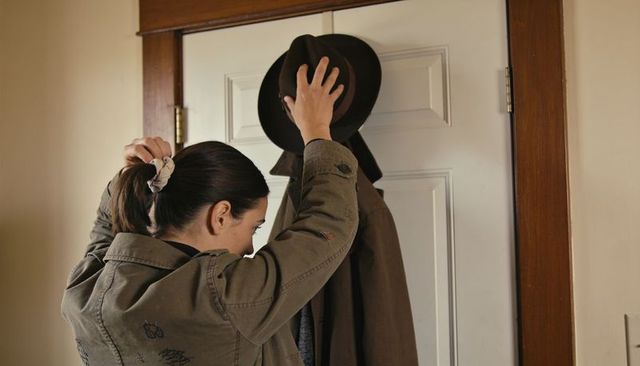 Woman reaching for fedora on door hook in cozy entryway with olive jacket