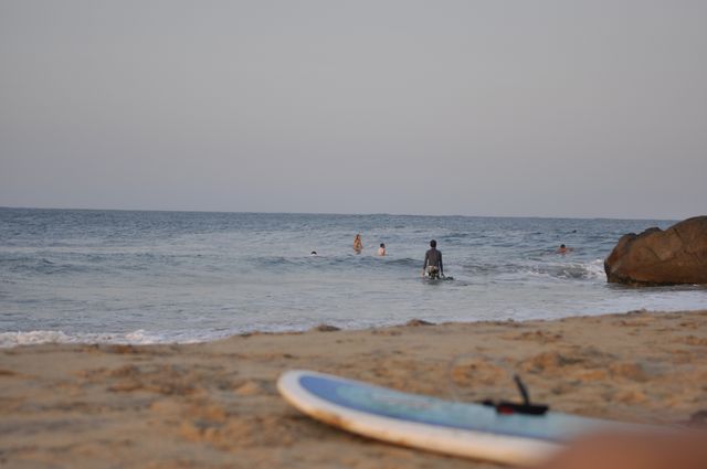 Several people seen enjoying the beach and swimming in the ocean during sunset. A surfboard is resting on the sandy shore in the foreground, providing a serene and relaxed atmosphere. This can be used for themes like vacations, relaxation, summer, and beach activities.