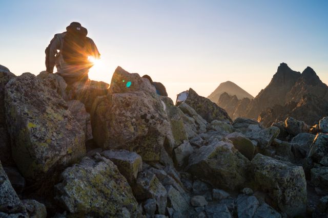 Person resting on rocky mountain summit as sun sets, creating tranquil silhouette. Ideal for promoting outdoor activities, adventure tourism, trekking equipment, nature contemplation, and wilderness resilience themes. Perfect for use in inspirational blogs, travel brochures, and adventure magazines.