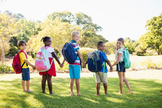 Diverse group of kids posing together in a park on a sunny day, each carrying backpacks. Ideal for use in educational materials, advertisements for children's products, or campaigns promoting diversity and inclusion.