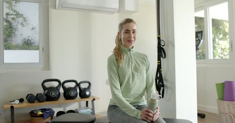 Young woman holding water bottle sitting on bench in bright home gym smiling near kettlebells