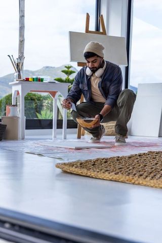 Young artist kneeling mixing paint with wooden bowl in bright studio by window
