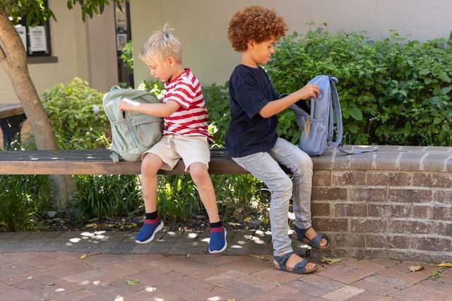 Two boys sorting backpacks on bench in shaded school courtyard back-to-school moment