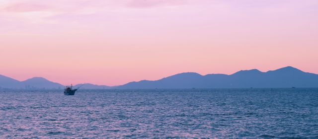 Serene scene features calm ocean waters during sunset, with a boat visible in the distance and mountains on the horizon. Soft light casts a pink hue across the sky, evoking tranquility and peace. Ideal for travel brochures, background images, and nature-themed projects.