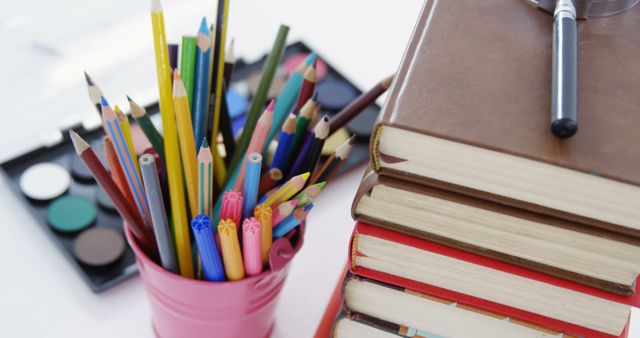 Colored pencils in pen holder with pile of books and magnifying glass against white background