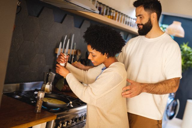 Biracial young man standing by smiling girlfriend spraying pepper on scrambled eggs in cooking pan. Unaltered, lifestyle, love, togetherness, food, preparation, beard, afro hair and home conept.