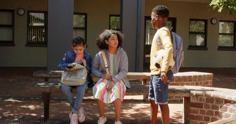 Diverse teens chatting on school courtyard bench with soccer ball and backpacks at school
