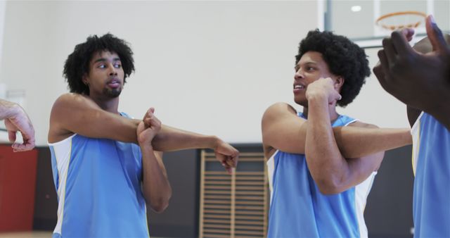Two male basketball players with afro hairstyles in blue uniforms are stretching their arms in preparation for a game on an indoor basketball court. This image can highlight activities on sports blogs, fitness training materials, team-building articles, and promotional content related to sports events or athletic gear.