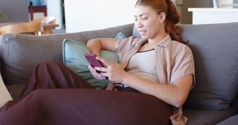 Relaxing woman texting on purple smartphone while reclining on cozy gray sofa