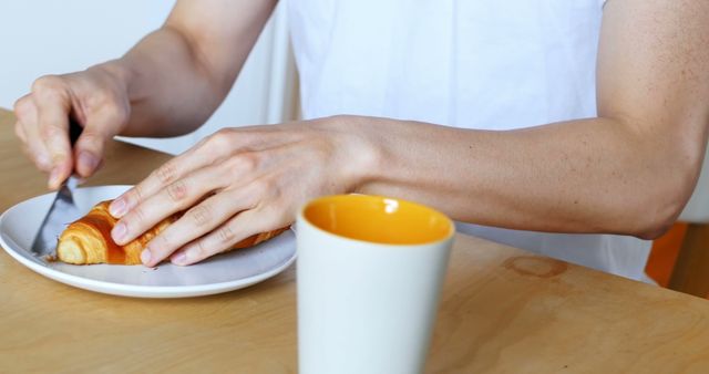 A person is slicing a croissant on a plate, with a cup of coffee nearby, with copy space. Capturing a simple breakfast moment, the image evokes a sense of morning routine and tranquility.
