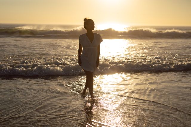 Woman walking along the beach at sunset with the ocean waves gently crashing in the background. Ideal for use in travel brochures, wellness blogs, relaxation and meditation content, and advertisements promoting beach vacations or serene getaways.
