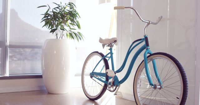 A vintage blue bicycle is parked indoors near a window with a view of a mountain, with copy space. Sunlight streams through the window, creating a warm, inviting atmosphere and highlighting the concept of eco-friendly transportation.
