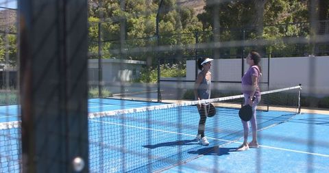 Mature women standing at net on blue paddle court holding paddles in sunlight
