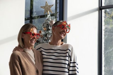 Smiling women wearing novelty christmas glasses by flocked tree, cozy holiday friendship
