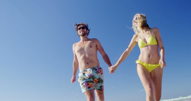 Happy couple holding hands while running on beach during summer vacation. Man wearing sunglasses and floral swim shorts. Woman in yellow bikini enjoying seaside. Perfect for promoting travel agencies, summer vacations, seaside resorts, and healthy lifestyle campaigns.