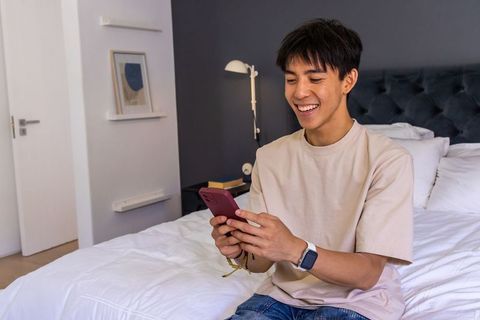 Young asian man smiling while texting on smartphone in modern minimalist bedroom