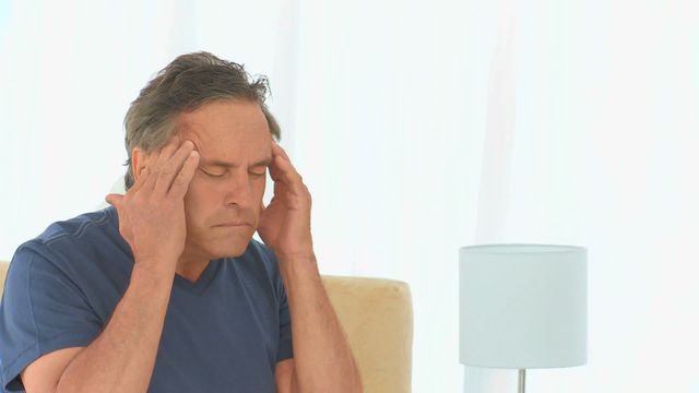 Middle-aged man sitting at home and holding his head in pain, suggesting headache or stress. Suitable for use in health and wellness contexts, articles on headaches, stress, and pain management tips. Also useful for illustrating symptoms of common ailments in medical and well-being contexts.