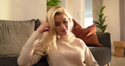 Blonde woman relaxing on sofa in cozy knit sweater with rust pillow and indoor plant