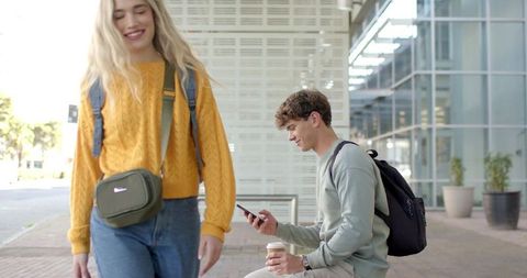 Students passing on college campus plaza, man using smartphone while holding coffee