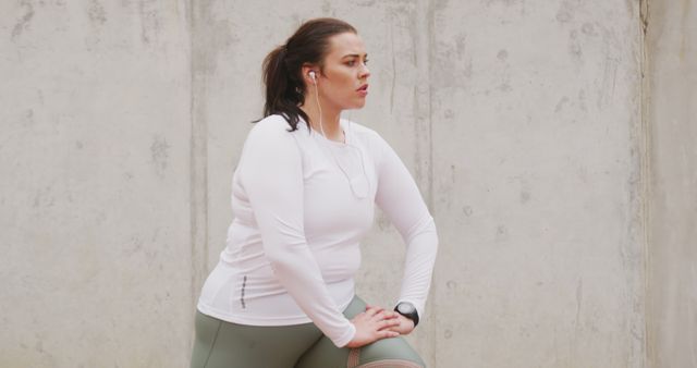 Woman pausing during outdoor workout, leaning on bent knee, wearing sportswear and earphones, and looking focused. Ideal for topics related to fitness, motivation, health, and outdoor activities.