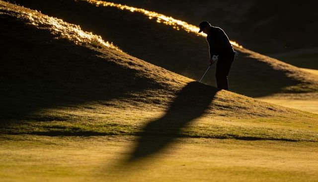 Golfer preparing chip shot on sunlit berm with dramatic long shadow at golden hour