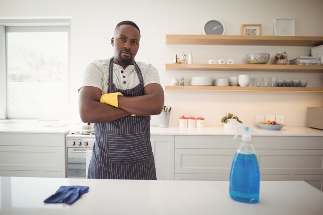 Confident man standing in a modern kitchen with arms crossed, wearing an apron and gloves. Cleaning supplies are visible on the counter. Ideal for use in articles or advertisements related to household chores, cleaning services, domestic life, and home hygiene.