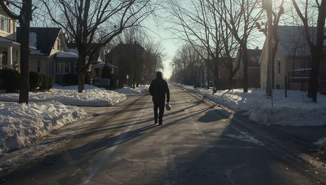 Walking silhouette on snowy suburban street at sunrise, hooded figure carrying bag