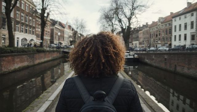 Curly-haired traveler walking canal path wearing backpack, historic european riverside