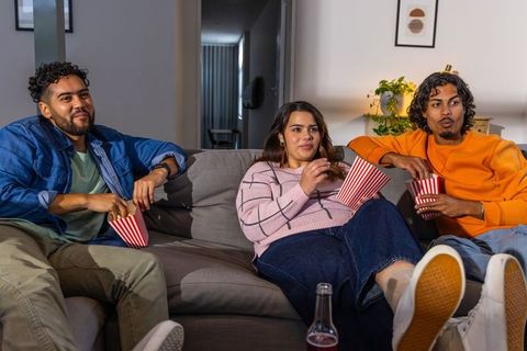 Multicultural friends watching movie on couch sharing popcorn, relaxing at home