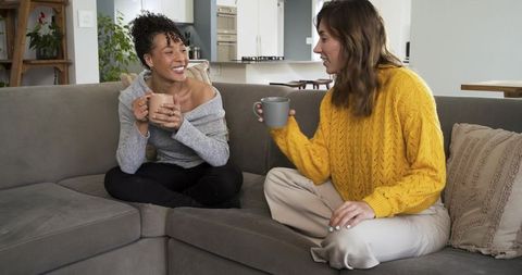 Multicultural friends sharing coffee, chatting on cozy gray sofa in modern kitchen