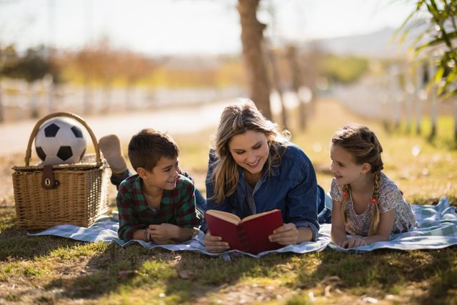 Mother reading a book to her children while lying on a blanket in a park on a sunny day. Ideal for use in family-oriented advertisements, educational materials, parenting blogs, and lifestyle articles promoting outdoor activities and family bonding.
