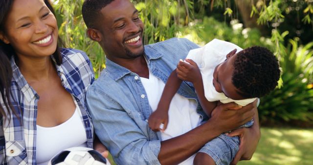 A young family is enjoying time together in a park. The mother is holding a soccer ball, and both parents are smiling while the father is playfully holding their child. This image can be used for promoting family-oriented products, outdoor activities, happiness, and bonding-related themes.