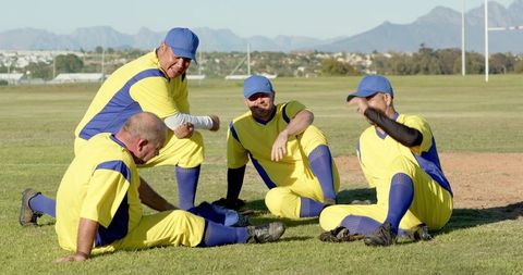 Veteran cricket team sitting and laughing on sunlit field in yellow and blue uniforms