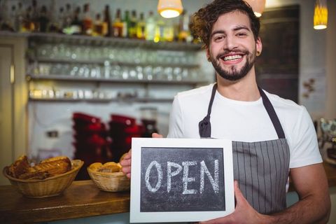 Cheerful Barista Holding Open Sign in Welcoming Cafe Environment