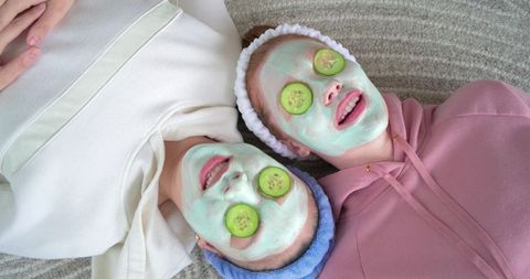 Two friends enjoying cozy home spa with green facial masks and cucumber slices
