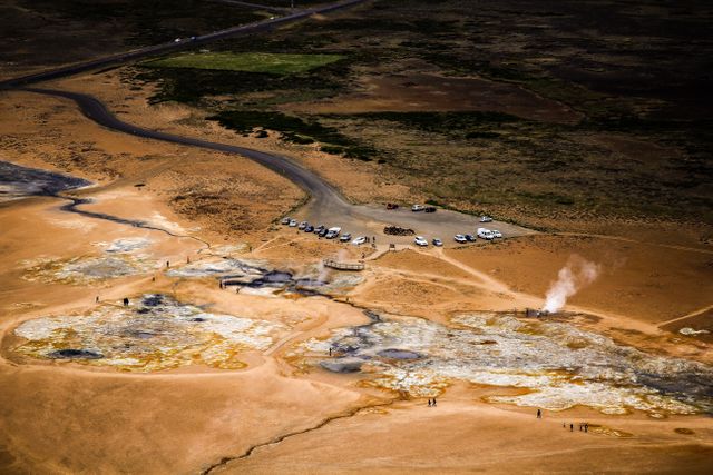 Aerial view capturing a geothermal area in a desert landscape. Visible are small groups of visitors exploring the steaming ground vents and parked cars indicating an attraction point. This image is suitable for use in articles or content about geothermal activity, natural wonders, travel destinations, or environmental studies.