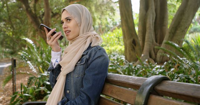 Young Muslim woman wearing a denim jacket and beige hijab, sitting at a park bench while using her smartphone's voice assistant feature. Greenery and trees surround her, creating a serene, natural environment. This image can be used for technology-related topics, outdoor leisure activities, or promoting voice assistant features among diverse communities.
