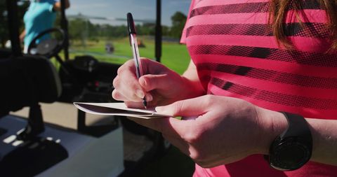 Caucasian woman in striped pink shirt writing in notebook at golf course