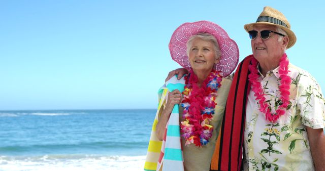 Elderly couple standing on a beach, smiling and wearing hats and leis. They look happy and content with brightly colored accessories like striped towels draped over their shoulders. The ocean is in the background with a clear blue sky overhead. Perfect for illustrating senior travel, leisure, families, happiness in retirement, tropical vacations, or lifestyle. This image can be used in senior wellness magazines, travel brochures, marketing campaigns targeted at older audiences, or retirement planning materials.