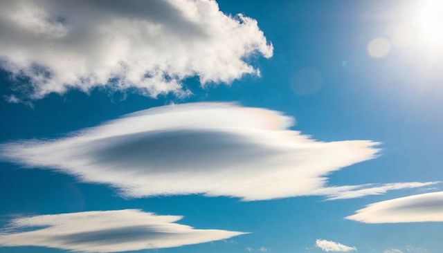 Lenticular cloud hovering over deep blue sky with sun lens flare and cumulus fragments