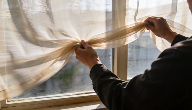 Hands pulling sheer beige curtain at sunlit window morning light soft drapery texture