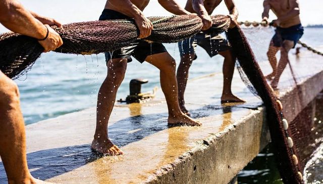 Shirtless fishermen hauling heavy wet net onto concrete pier with buoys, coastal teamwork