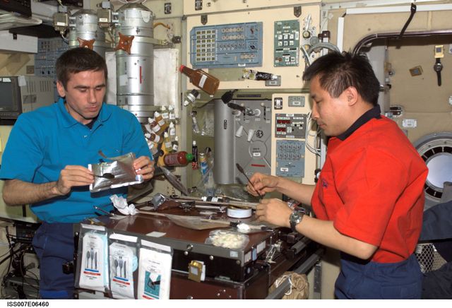 Astronauts in ISS Zvezda Service Module sharing a meal while conducting space missions. Can be used for themes related to space exploration, teamwork, or life in space.