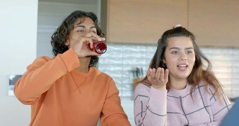 Friends chatting at kitchen counter drinking red beverage while using laptop for call