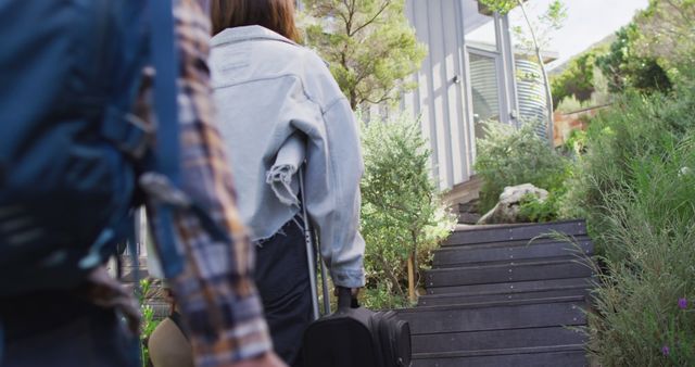 Two travelers are walking up the stairs towards a modern house, carrying luggage and backpacks. They are surrounded by greenery and appearing excited to reach their destination. This can be used in travel-related articles, vacation home rentals promotion and lifestyle blogs focusing on adventure and holiday themes.